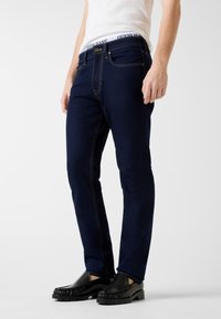 Jeans di denim blu navy con vestibilità slim, cuciture gialle a contrasto e cintura con logo. Indossati con scarpe slip-on nere.