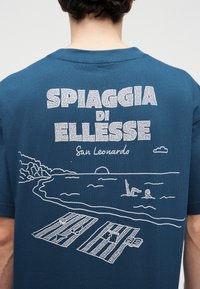 Πίσω μέρος μπλε πουκάμισου με άσπρη γραμμική τέχνη παραθαλάσσιας σκηνής, δύο άνθρωποι κολυμπούν, ο ήλιος δύει, καρέκλες παραλίας και το κείμενο «SPIAGGIA DI ELLESSE San Leonardo».
