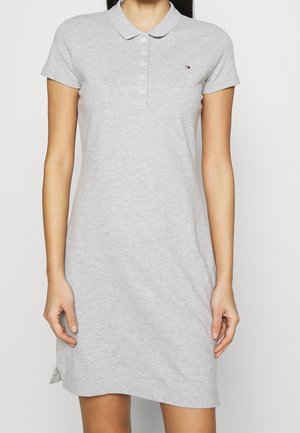 Mujer vistiendo un vestido tipo polo de manga corta, gris claro, con un pequeño logo bordado en el pecho.