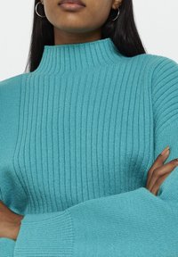 Tyrkysový pletený svetr s vysokým límcem a širokými rukávy. Měkká textura a rovnoměrné stehy po celé látce.