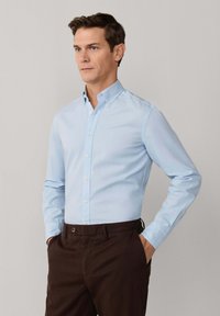 Mężczyzna w jasnoniebieskiej koszuli z guzikami i ciemnobrązowych spodniach, stojący z rękami w kieszeniach, patrzący przed siebie.