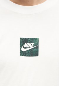 Κοντινό πλάνο ενός λευκού πουκαμίσου με ένα πράσινο τετράγωνο που φέρει το λευκό λογότυπο Nike και το σήμα swoosh στο κέντρο του στήθους.