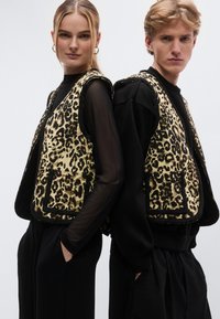 Bézs és fekete leopárdmintás mellények, puha tapintásúak és széles karöltőkkel. Fekete hosszú ujjú felsők és nadrágok fölé viselve.