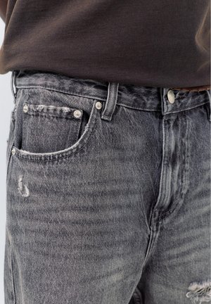 Záber zblízka na vyblednuté čierne denimové džíny s malými opotrebovaniami pri prednom vrecku, nosené s hnedou košeľou zastrčenou vo vnútri a hnedým opaskom.