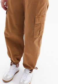 Bruine cargobroek met elastische boorden en zijzakken, gemaakt van een gladde, lichtgewicht stof; gecombineerd met witte sport sneakers.