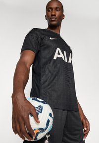 Maillot de football Nike noir avec un design texturé, arborant "AIA" en blanc. Le modèle tient un ballon de football à motif avec des accents bleus et oranges.