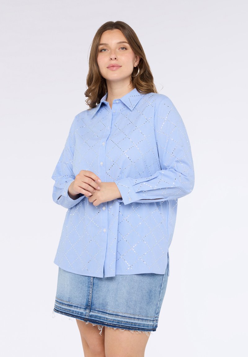 Camicia button-up azzurra con ritagli a forma di diamante, maniche lunghe e colletto classico, abbinata a una gonna di jeans con orlo sfrangiato.