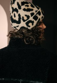 Zwarte gestructureerde jas met een hoge kraag, gedragen met een crèmewitte beanie met een zwart luipaardpatroon. Krullend haar is gedeeltelijk zichtbaar.