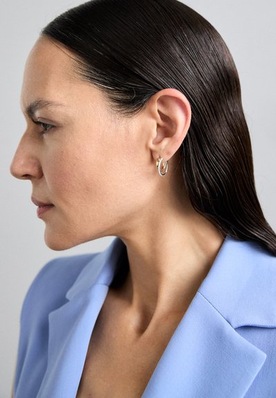 Aros en oro y plata llevados por una mujer con cabello oscuro y liso, vistiendo un blazer azul claro, frente a un fondo neutro.