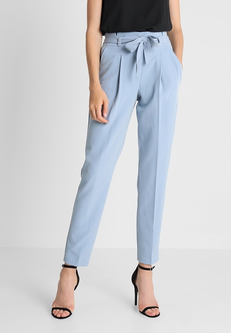 Femme portant un pantalon taille haute bleu clair avec ceinture nouée, haut noir sans manches et sandales noires à talons hauts, debout devant un fond blanc.
