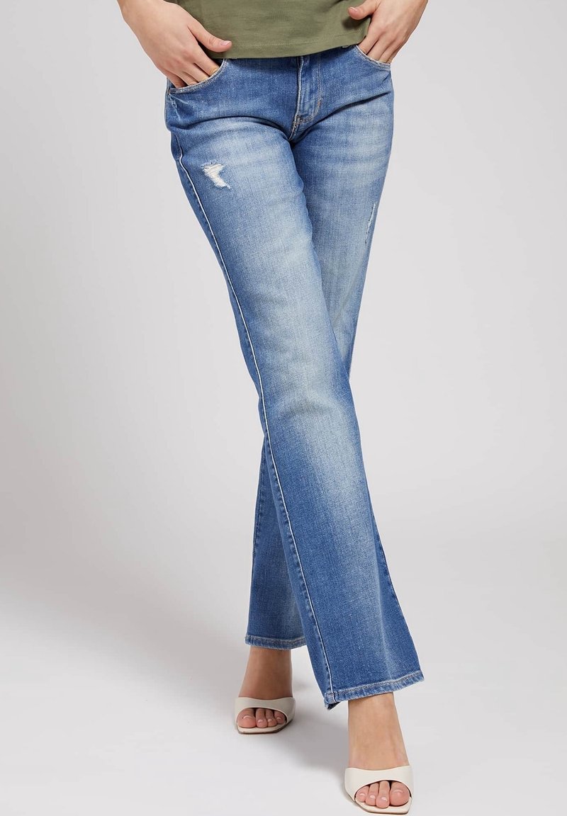 Personne portant un jean droit bleu clair déchiré et des talons ouverts beige, debout les mains dans les poches sur un fond uni.
