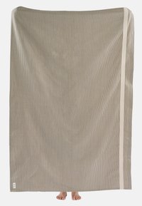 Pehme puuvillane rätik beežis toonis, millel on tekstuurne mustriga disain ja üks serv on kaunistatud kreemja triibuga. Alumises osas on näha jalgu.