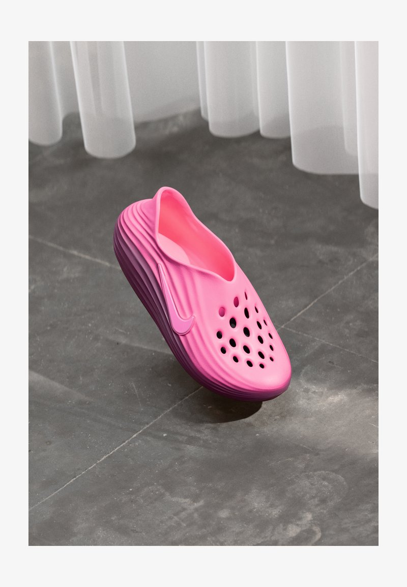 Clog rosa brilhante e perfurado, com sola texturizada, repousando sobre um chão de pedra cinza sob cortinas brancas transparentes.