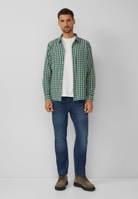 Man staat in een groen-wit geruit overhemd over een wit T-shirt, blauwe jeans en bruine veterschoenen voor een effen grijze achtergrond.