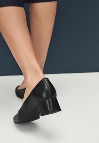 Le scarpe nere in pelle con tacco a blocco presentano una superficie liscia, una punta arrotondata e un sottile accento dorato sul tacco come dettaglio.