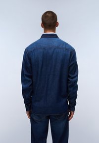 Hombre de pie de espaldas, vistiendo una chaqueta de mezclilla azul oscura y jeans a juego, frente a un fondo gris claro.