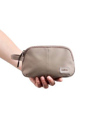 Main tenant une petite pochette taupe avec fermeture éclair, boucle en tissu et étiquette "bellroy" sur le devant, sur fond blanc.