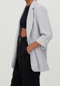 Femme portant un blazer gris clair aux manches retroussées, un crop top noir, un pantalon taille haute noir et une ceinture noire avec une grande boucle.