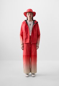 Red waterproof jacket with gray accents, paired with matching loose pants featuring an ombre effect. Accompanied by a red wide-brimmed hat.