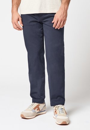 Homme portant un pantalon bleu marine et des baskets beige avec des accents marron, debout devant un fond blanc uni.