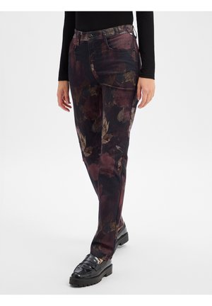 Person trägt dunkel gemusterte, eng anliegende Hose mit Blumenmuster und schwarze Loafers, steht vor einem einfarbigen hellen Hintergrund.