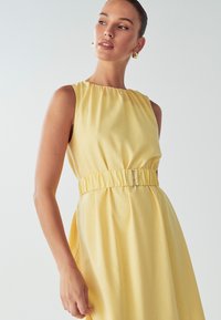 Keltainen hihaton mekko, jossa on poimutettu kaula-aukko ja vyötärö. Mekossa on joustava vyötärö, jossa on ohut metallinen solki, sekä pehmeä kangas.