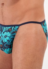 Schwimmslips mit einem blauen Blumenmuster auf dunklem Hintergrund, bestehend aus einem glatten Material und einem schmalen Bund.