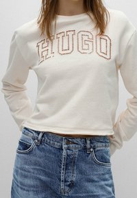 Sudadera recortada de color crema con el diseño recortado de "HUGO". Mangas largas y un corte relajado, combinada con jeans de mezclilla azules.