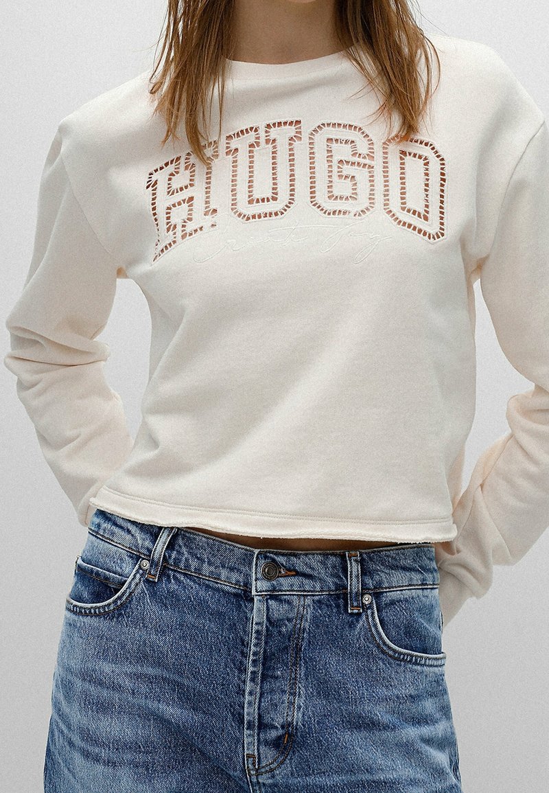 Sudadera recortada de color crema con el diseño recortado de "HUGO". Mangas largas y un corte relajado, combinada con jeans de mezclilla azules.