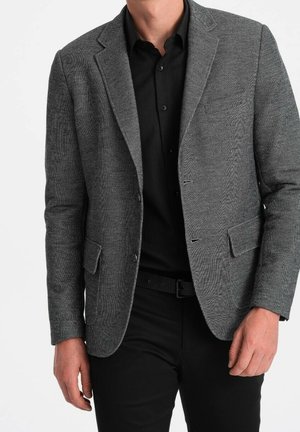 Hombre con blazer gris texturizado sobre una camisa negra de botones y pantalones negros, de pie con las manos relajadas a los costados.