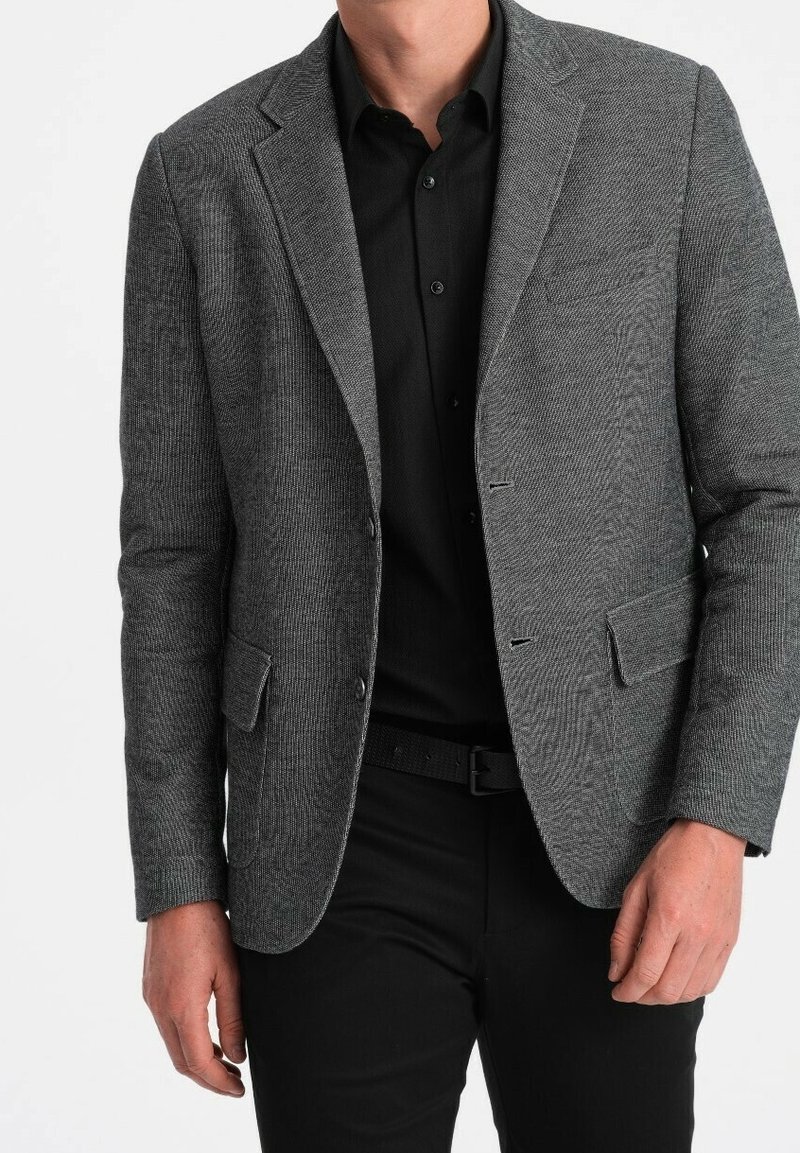 Homme portant un blazer gris texturé par-dessus une chemise noire boutonnée et un pantalon noir, debout les mains détendues le long du corps.
