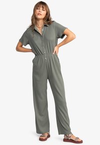 Olijfgroen geribd jumpsuit met korte mouwen, knopen aan de voorkant en een cinched taille. Model draagt kleurrijke sandalen. Volledige lengte, relaxte pasvorm.