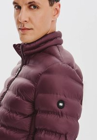 Śliwkowa kurtka puchowa z wysokim kołnierzem, zapięciem na zamek i okrągłą czarną naszywką z logo na lewym ramieniu, z pikowanymi sekcjami i gładką teksturą.