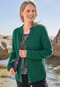 Groene textuur cardigan met een ronde hals en rits, gedragen over een gestreept topje. Bevat een voorkantzak en heeft een losse pasvorm.