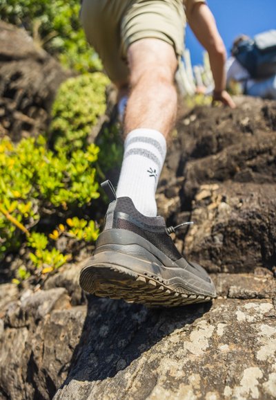 Svarta vandringsskor med mesh-överkäng, gummisula och texturerat mönster, visade på stenig terräng. Vita strumpor med grå ränder synliga.