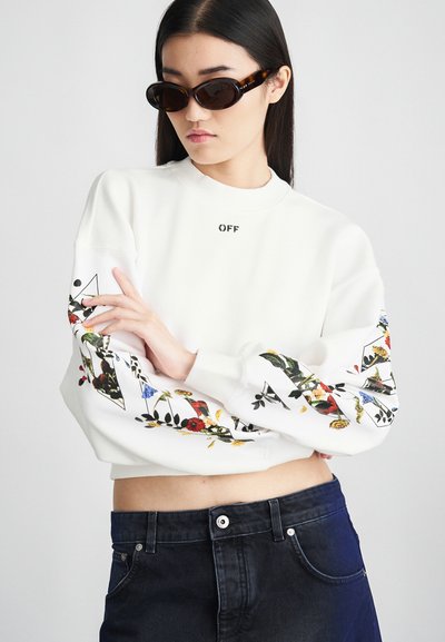 Sweatshirt blanc avec des motifs floraux sur les manches, col rond et logo "OFF" sur la poitrine. Le modèle porte un jean sombre et des lunettes de soleil marron.