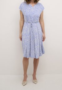 Robe à motifs bleus et blancs avec manches courtes, col en V, ceinture à la taille et jupe évasée. Portée avec des chaussures beige clair.