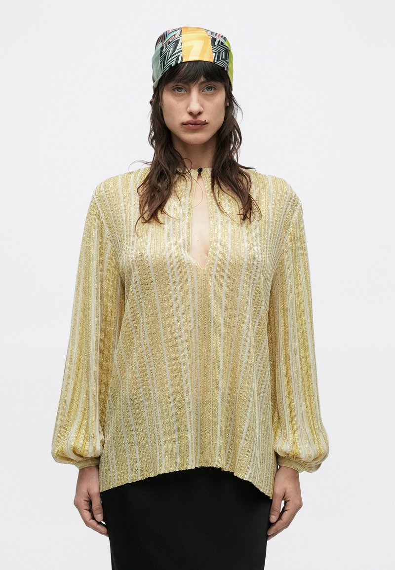 Kvinde iført en gulstribet bluse med lange ærmer og nøglehulskæring samt et flerfarvet mønstret tørklæde, stående mod en lys baggrund.