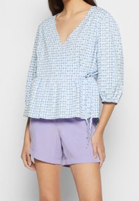 Jasnoniebieski top peplum w kratę z dekoltem w serek, bufiastymi rękawami i detalem ze sznurkiem, w komplecie z lawendowymi szortami. Gładka faktura tkaniny.