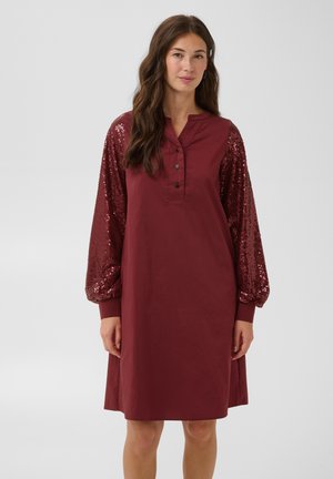 Femme portant une robe bordeaux longueur genou avec des manches longues couvertes de sequins et un col boutonné, debout devant un fond uni.