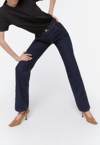 Mørkeblå lige ben jeans lavet af denim, med kontrasterende orange syninger og frontlommer. Sammen med camelfarvede højhælede sko.