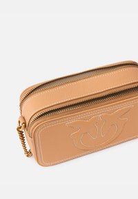 Borsetta in pelle tan con doppio zip, design embossato, cuciture bianche e finitura testurizzata. Presenta un accento di catena dorata su un lato.