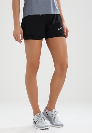 Female wearing black Nike shorts and gray and white Nike running shoes, standing on a plain light background.