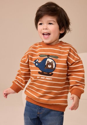Tout-petit souriant portant un sweat-shirt rayé orange avec un patch hélicoptère bleu et ours, associé à un jean bleu, debout sur un fond neutre.