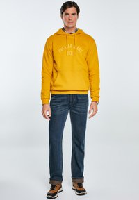 Żółta bluza z kapturem i kieszonką kangurką, z sznurkiem do regulacji, z napisem "BIG STAR JEANS 1979". W połączeniu z ciemnoniebieskimi dżinsami i brązowymi trampkami.