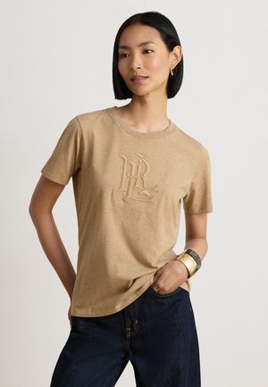 Femme aux cheveux courts noirs portant un t-shirt beige à manches courtes avec un monogramme en relief et un jean bleu foncé, posant avec une main dans la poche et des bracelets en or.
