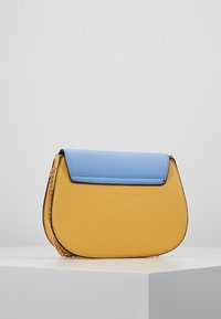 Bolso con un cuerpo amarillo y una solapa azul, hecho de un material suave. Presenta un acento de cadena en el costado y una forma redondeada.