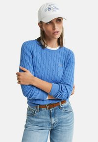 Un pull en jersey bleu clair avec des poignets côtelés, porté par-dessus une chemise blanche. Assorti avec un jean bleu clair et une casquette de baseball blanche avec un texte bleu.