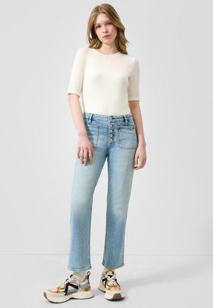 Vrouw staat met een wit shirt met korte mouwen, lichtblauwe high-waisted jeans met knopen aan de voorkant en witte sneakers met oranje en zwarte accenten.