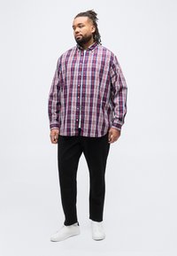 Άνδρας με dreadlocks που φοράει κόκκινο, λευκό και μπλε καρό πουκάμισο με κουμπιά, μαύρο παντελόνι και λευκά αθλητικά παπούτσια, στέκεται μπροστά σε απλό φόντο.